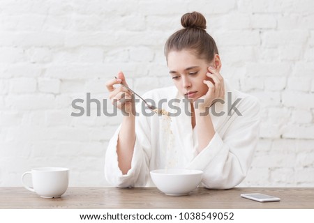 Young woman with no appetite, tired, bad mood and sleepy, sitting in the white loft kitchen, having unsavory unpalatable unappetizing breakfast