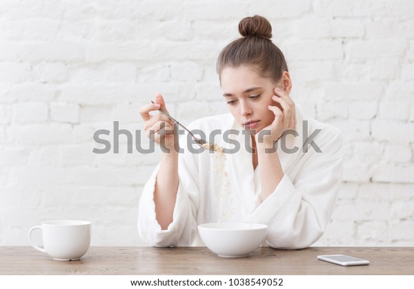 Young woman with no appetite, tired, bad mood\
and sleepy, sitting in the white loft kitchen, having unsavory\
unpalatable unappetizing\
breakfast