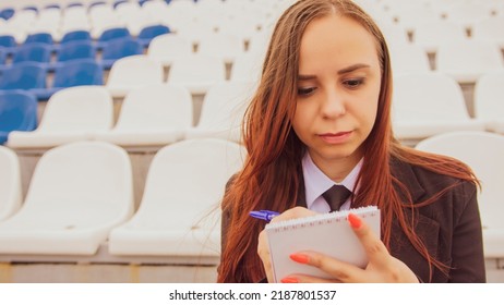 Young Woman With Notepad, Pen Sitting On Stadium Bleachers Alone. Female Journalist Writing Down Notes During Sports Training At Street Stadium