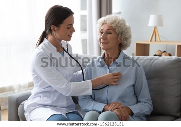 Young woman nurse doctor gp holding stethoscope\
examining old senior 60s grandma patient check heartbeat at\
homecare checkup medical visit at home hospital, older people\
cardiology healthcare\
concept
