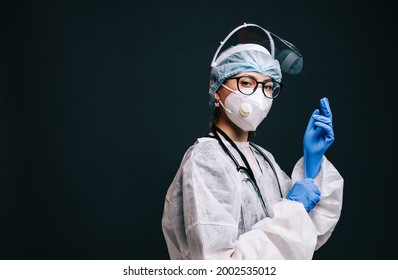 Young Woman Nurse Hospital Worker In Medical Protective Mask, Gloves And Protective Wear, Ready For Work. Isolated On Dark Background With Copy Space.