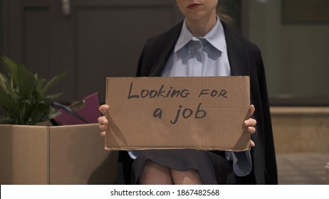 Young woman in office clothes with a sign looking for a job sitting with cardboard box. Woman in office skirt looking for a job on the street, no face