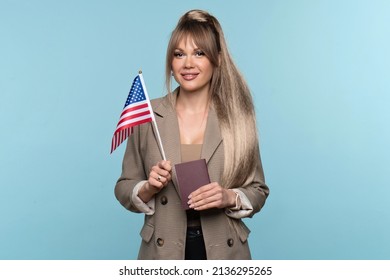 Young Woman In Office Suit Showing Small Flag USA And Passport On Light Blue Backdrop. Study Abroad Concept. International Student Exchange Program. Learning American English With Native Speaker