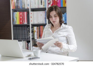 Young Woman In Office Working On Desktop. She Is Reviewing Papers.