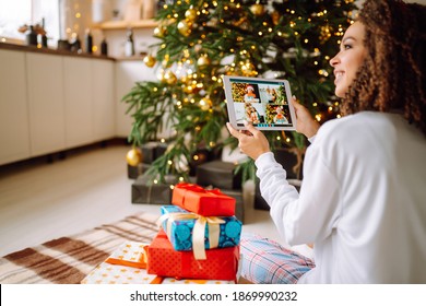 A Young Woman On The Background Of A Christmas Tree With Gifts, With A Tablet Has A Video Call Or Video Chat With Friends, Family. Christmas Online Holiday. Stay Home Vacation.	