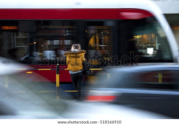 Young woman on a bus stop on city street,\
cars and trams passing by in motion\
blur