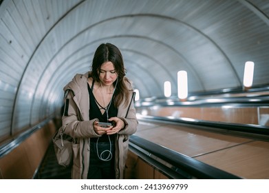 A young woman on an escalator in the subway with a phone in her hands. A beautiful girl rises from the subway on an escalator, listens to music with headphones, holds a cell phone - Powered by Shutterstock