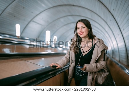 A young woman on an escalator in the subway with a phone in her hands. A beautiful girl rises from the subway on an escalator, listens to music with headphones, holds a cell phone