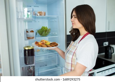 Young Woman On Kitchen During Quarantine. Stand In Front Opened Fridge And Hold Plate With Fruit, Citrus And Cookies. Ready To Cook.