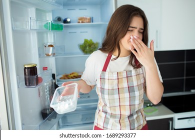 Young Woman On Kitchen During Quarantine. Hold Plasti Foot Tray Opened With Bed Smell. Unfresh Food. Woman Feel Sick Because Of Bed Smell. Stand In Front Of Opened Fridge.