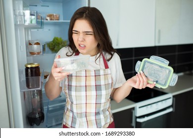 Young Woman On Kitchen During Quarantine. Stand In Front Opened Fridge With Food Tray With Bad Smell. Disgution.