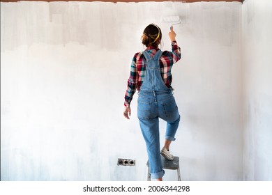 Young Woman On A Ladder Painting New Apartment Walls In Color White With Paint Roller. Renovating House. Real Estate Concept. Copy Space