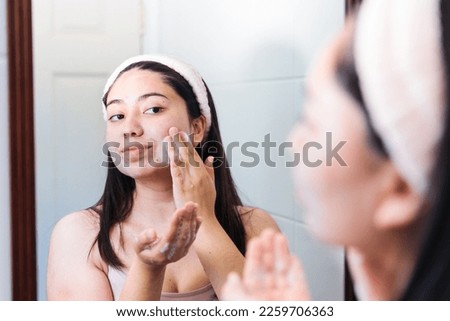 Young woman on the mirror, applying hydrating cleanser soap on her face. Skin care at home.
