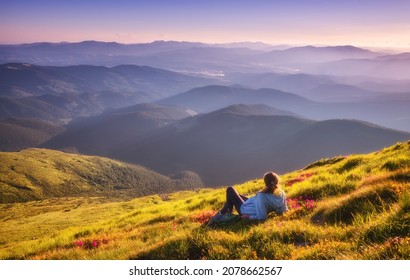 Young Woman On The Mountain Peak With Green Grass And Pink Flowers Looking At Beautiful Mountains In Fog At Sunset In Summer. Colorful Landscape With Lying Girl, Forest, Hills, Sky. Travel And Tourism