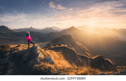 Young woman on mountain peak and beautiful mountain valley in haze at colorful sunset in autumn. Dolomites, Italy. Sporty girl, mountain ridges in fog, orange grass, trees, golden sun in fall. Hiking	