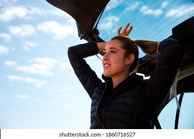 Young Woman On Road Trip Sitting In Open Tailgate Of Car Putting Hair In Pony Tail Before Heading Off On Hike In Countryside Against Blue Sky