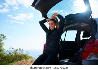 Young Woman On Road Trip Looking Into Camera And Smiling As She Sits In Open Tailgate Of Car Putting Hair In Pony Tail Before Heading Off On Hike In Countryside Against Blue Sky