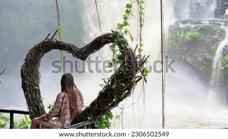a young woman on a swing in the shape of a heart, enjoying the rainforest and looking at the high powerful waterfall Tegenungan. Rear view of a tourist girl on the Indonesian island of Bali