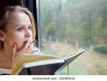 Young Woman On A Train Writing Notes In A Diary Or Journal Staring Thoughtfully Out Of The Window With Her Pen To Her Lips As She Thinks Of What To Write