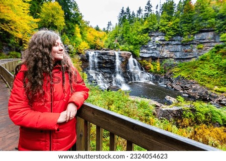 Young woman on wooden boardwalk deck overlook railing at Blackwater Falls waterfall state park in West Virginia autumn fall season looking at view happy