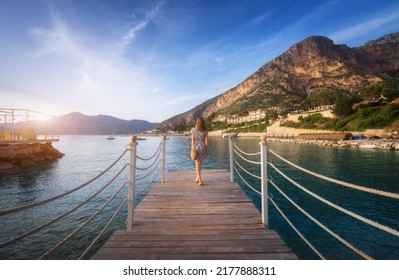 Young woman on wooden pier on sea shore is looking on mountain, blue water and sky at sunset in summer. Girl on jetty, beach, hotels on the hill, green trees, city, rock, boats in Oludeniz, Turkey