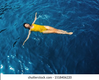 young woman in a one-piece yellow swimsuit floats on the surface of the water. Brunette girl relaxes and bathes in the blue sea, relaxing by the sea