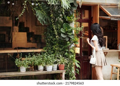 Young woman opening door of modern cozy coffeeshop with walls decorated with lush plants