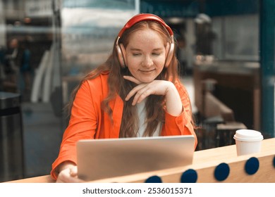 A young woman in an orange blazer enjoys music while working on her laptop in a cozy café on a sunny day - Powered by Shutterstock