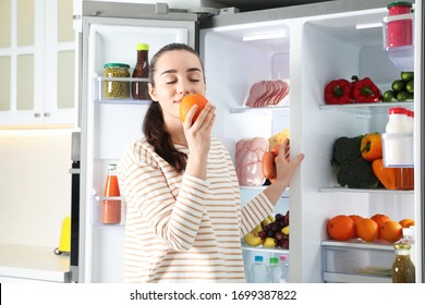 Young woman with orange near open refrigerator indoors