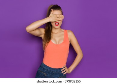 Young Woman In Orange Shirt Cover Eyes.Waist Up Studio Shot On Purple Background.