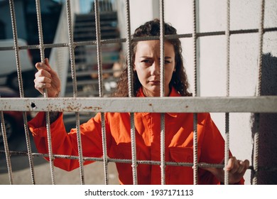 Young woman in orange suit behind jail bars. Female in colorful overalls portrait. Law and justice concept.
