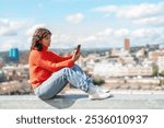 A young woman in an orange sweater sits on a ledge, enjoying the skyline and using her phone on a sunny day