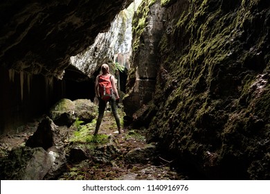 Young woman with orange travel backpack and lightern explores ancient fortress cave, view from back