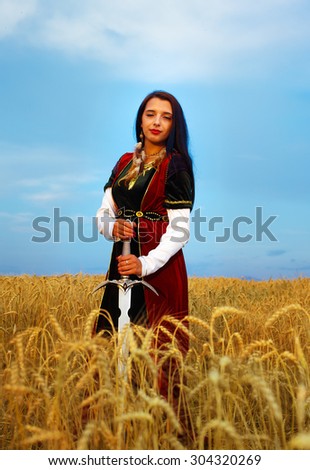 Young woman with ornamental dress and sword in hand  standing on a wheat field with sunset. Natural background.