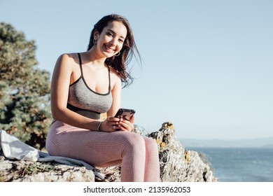 Young Woman Outdoors On Sport Clothes Checking Her Smart Phone After Exercise. Carefree, Positive And Freedom Concept. Healthy Lifestyle Doing Exercise Outdoors. Super Smile With Confident Attitude