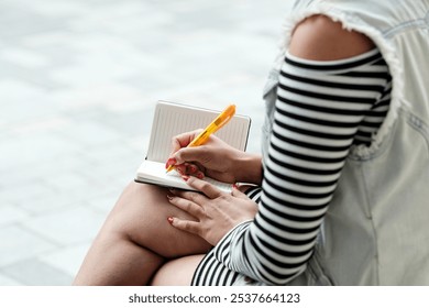 Young woman outdoors, writing notes in a notebook, taking a break in the city, casual and creative in the sunlight - Powered by Shutterstock