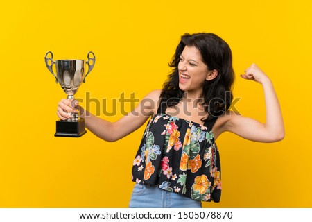 Young woman over isolated yellow background holding a trophy