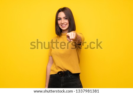 Young woman over yellow wall points finger at you with a confident expression