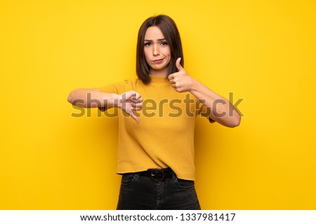 Young woman over yellow wall making good-bad sign. Undecided between yes or not