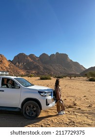 Young Woman In Overalls With Hat Stands Near Car In Desert. SUV Offroad Auto Vehicle White With Camping Equipment And Rooftop Tent. Safari Travel. Spitzkoppe Mountain In Africa, Namibia.