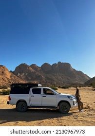 Young Woman In Overalls With Hat Stands Near Car In Desert. SUV Offroad Auto Vehicle White With Camping Equipment And Rooftop Tent. Safari Travel. Spitzkoppe Mountain In Africa, Namibia.