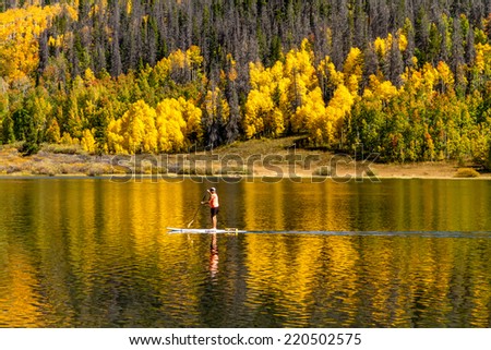 Young woman paddle boarding on mountain lake on warm fall day with yellow Aspen trees reflecting in water