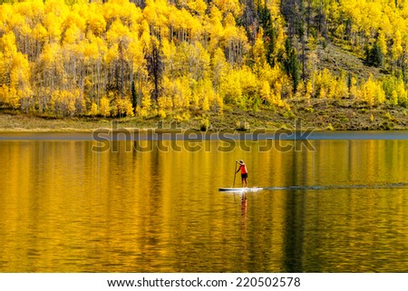 Young woman paddle boarding on mountain lake on warm fall day with yellow Aspen trees reflecting in water