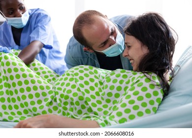 Young Woman In Pain Pushing For Childbirth At Maternity While African American Nurse Assisting In Hospital Ward. Person Giving Birth While Laying In Bed And Father Of Baby Holding Hand