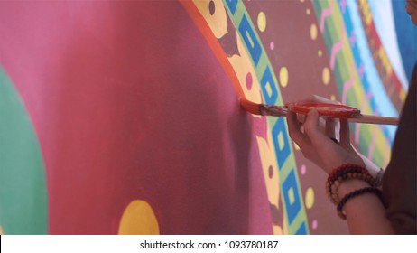 Young Woman Painting Graffiti Or Mural On Wall With Brush, Close-up. Clip. Painter Performing Street Art. Close-up On Hands Of A Painter With A Brush In Acrylic Paint