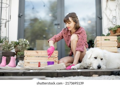 Young Woman Painting Wooden Box In Pink Color, Doing Housework While Sitting With Her Dog On Terrace Of Her House. DIY Concept. Idea Of Friendship With Pets