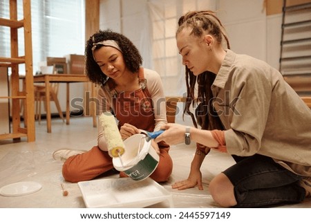 Young woman with paintroller sitting on the floor next to African American female colleague in workwear pouring white paint in plastic tray