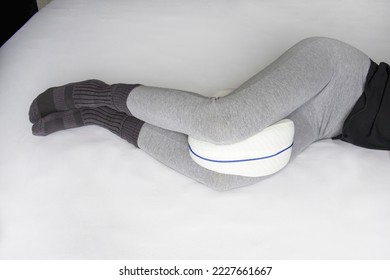 Young woman in pajama pants with an anatomical pillow between her legs and knees, lying on a bed with white sheets