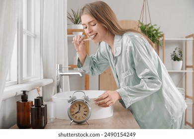 Young woman in pajamas with alarm clock brushing teeth at home - Powered by Shutterstock