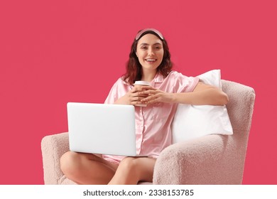 Young woman in pajamas with cup of coffee using laptop in armchair on pink background - Powered by Shutterstock